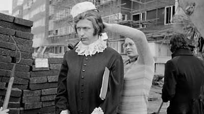 Graham Chapman is dressed as an Victorian old lady by a costumier. He is smoking a pipe