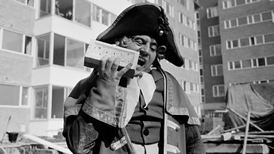 Terry Jones dressed as the Dickensian character Mr Bumble in large 'Nelson' hat. On a building site, he appears to be chewing a brick.