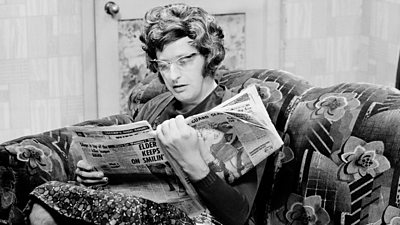 Graham Chapman dressed as a woman, sat on a floral sofa reading a tabloid newspaper. He is wearing very 1960s horn-rimmed glasses.