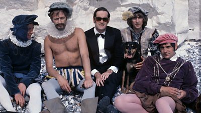 Michael Palin, Graham Chapman, John Cleese, Eric Idle and Terry Jones on a pebbly beach. All but Cleese are in Tudor minstrel outfits, Cleese is in black tie.