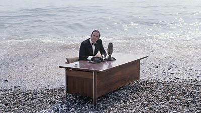 John Cleese, in full evening dress, sat at a news announcer's desk on a beach. The tide is coming in