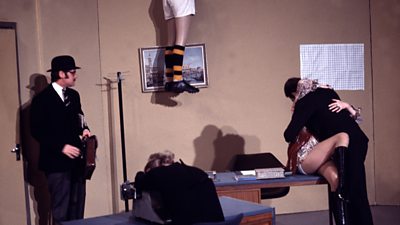 Michael Palin, dressed in suit and bowler hat, watches Carol Cleveland and John Cleese embracing passionately on a desk. A pair of legs in sportswear are dangling from the ceiling.