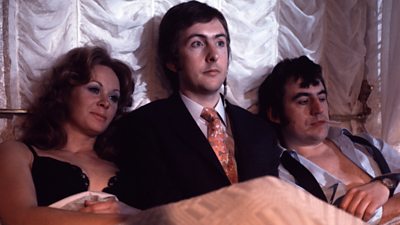 Eric Idle in a full suit looking sheepish between Carol Cleveland and Terry Jones in a bed, with the other two in a state of sultry undress.