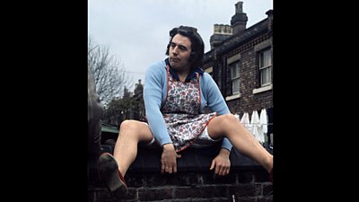 Terry Jones in drag with bri-nylon cardigan and hair in curlers sits with legs wide open on the yard wall of a row of terrace houses. 