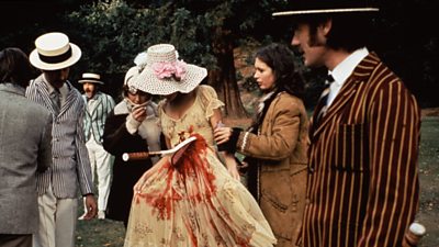 Carol Cleveland in Edwardian dress, has a tennis racket lodged bloodily into her abdomen. A costume assistant makes adjustments. Michael Palin and Eric Idle in straw boaters and stripy blazers look on. 