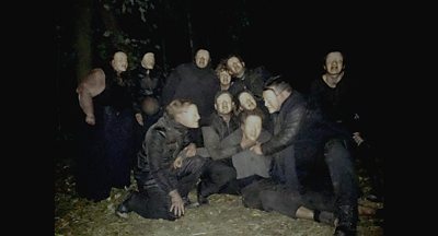 In a dark wood, a nightmarish masked mob surround a man