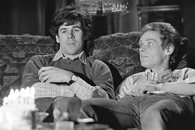 Two men on a sofa, with a birthday cake lit