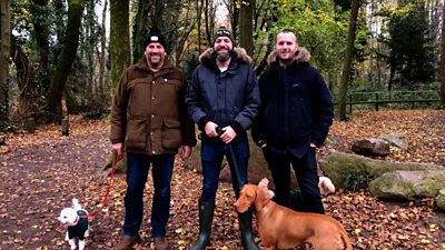 Three men looking into the camera on a dog walk with three dogs in the woods