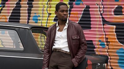 A young black man leans on a car holding a pistol. The shutter behind is covered in graffiti. 