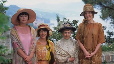 Four women in 1920s dress and hats line up smiling at the camera. A beautiful Italian valley is behind