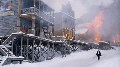 A lone man away from a burning building along the snow-covered street of a wooden gold prospecting town.