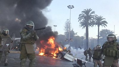 Riot police walk past a burning car