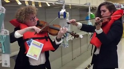 Two women playing violin in supermarket wearing lifejackets