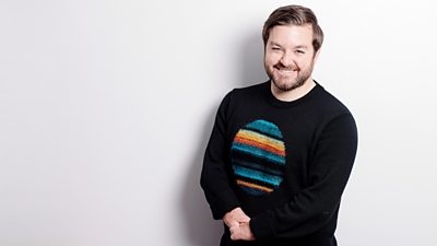 A smiling Alex Brooker mid-shot standing in front of a blank white wall, wearing a black jumper with a large circle on it that's filled with stripes of different orange, blue and black stripes.