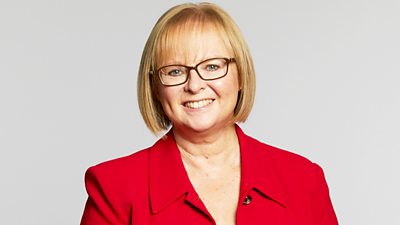 A headshot of a woman in a red collared jacket and black rimmed glasses smiling at the camera 