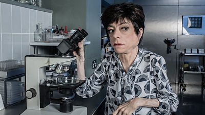 Liz Carr holding a microscope and looking right of camera