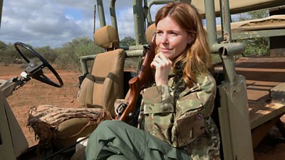 Stacey Dooley sitting in a jeep holding a rifle