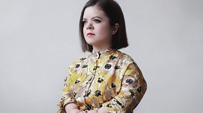 Sinéad Burke mid shot on a white background, wearing a floral dress, has a bob hair cut and not smiling. Looks deep in thought,