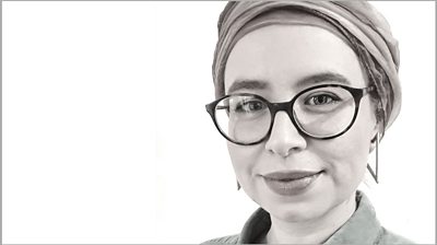 Black and white image of Erika Jones who is to the right of centre looking straight into camera, She's wearing a headscarf and glasses. 