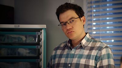 Jules Robertson as Jason Haynes in Holby City. Wearing a wide checked shirt which is white, grey and blue. Had glasses on and dark brown hair. In background are shelves with medical equipment