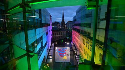 The exterior of New Broadcasting House lit up in the election colours. A huge physical map of the UK in hexagonal squares is in the middle of the piazza.