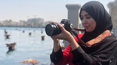 An El Kul journalist holds a camera preparing to take a photo beside water.
