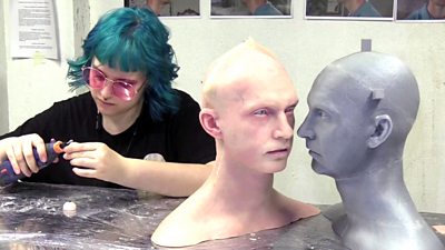 A woman works on a robot eye, next to her are two robotic heads