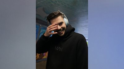 Syed Abbas Naqvi stands in an subway with graffiti behind him. Three-quarter length shot and he's smiling to camera, while rubbing his right eye. He's wearing a black hoodie with MAN embossed on the front and chunky grey headphones.