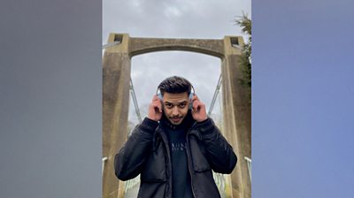 Syed Abbas Naqvi stands with a concrete looking bridge behind him. Three-quarter length shot and he looks straight to camera with both hands on his grey chunk headphones he has on his head. He's wearing a black hoodie with MAN embossed on the front and black jacket.