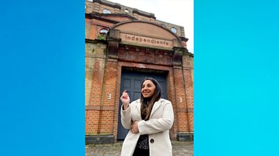 Shani Dhanda stands in front of a red brick building that looks quite industrial. She's shot at more than three-quarters. The photography attempts to contrast Shani's small stature with the enormity of the building behind.