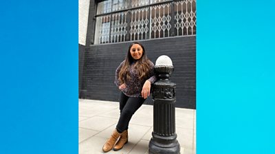 Shani Dhanda leans on a car bollard with her left arm and her legs crossed. She's shot in full-length. The photographer attempts to demonstrate Shani's small stature through contrasting the height of the bollard.