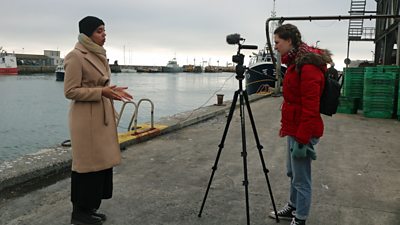 Star McFarlane stands near Plymouth Harbour facing camera on tripod wearing a black beanie and beige three-quarter length coat. Maxine Denton behind the camera in a red coat with rucksack