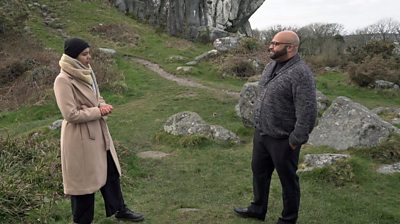 Star McFarlane stands in a green rocky area wearing a black beanie and beige three-quarter length coat. She is interviewing conductor Marcus Alleyne who is wearing a grey cardigan and charcoal trousers