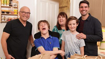 Presenter Greg Wallace (left) wearing a black t-shirt and smiling to camera, while standing next to a family of four (one mum and three boys) in a kitchen with Eat Well For Less food bags in front of them.