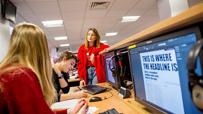 A male and female student sitting at computers editing an online website page which says "This is where the headline is", while another woman stands looking at them.