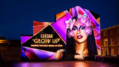 Shows a 3D mural at night time on the side of a building in Liverpool. Maya Jama on the right with purple make-up and flowers. Left tv Glow Up in gold, with Finding The Next Make-Up star below on a black background. Shades of purple and pink fill the rest of the space. Lit up with spot lights.