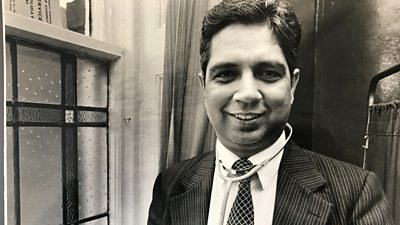 Black and white image of Professor Dr Shiv Pande midshot with a stained glass window of a cross to his right