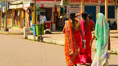 Women in India walking on a street in orange, pick and blue sarees. We are seeing them from the back - so no faces.