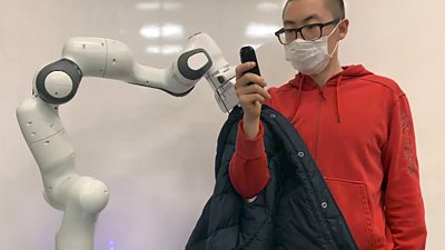 A robot helps a scientist who is wearing a red zip-up hoodie put on a black jacket