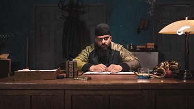 From Man like mobeen - with mobeen behind a heavy wooden desk, looking to camera. Wearing a beanie and bomber jacket