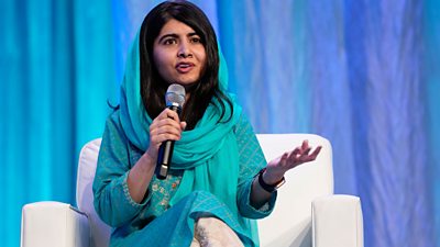 Malala Yousafzai sitting in a white seat, with a microphone in her right hand, holding out her left as she speaks. Background a blue curtain and she wears a turquoise salwar