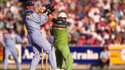 Javed Miandad bats wearing the Pakistan uniform - green with blue and red strips. He's in the background. In the foreground is a wicket keeper from the other team - wearing whites.