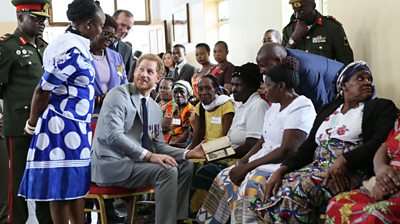 Prince Harry in the centre of image in light grey suit and black tie with medal on left breast. Around him are about a dozen Zambian men and women.