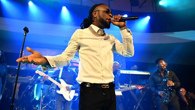 Burna Boy holding microphone to mouth with left hand, right hand is out to side. White shirt, black trousers. Behind him, the band. A man with a sax is visible.