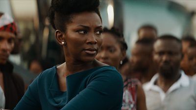 Genevieve Nnaji in the cetnre looking to her left. Tight mid-shot. Wearing green. Behind her out of focus around a dozen people.