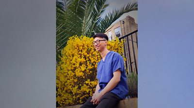 On a grey backing to make the image 16 x 9. Portrait of Dr David Lau in his NHS scrubs, wearing black rimmed glasses and smiling. Almost a side profile. Next to a yellow flowering bush.