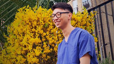 Dr Lau close up, with scrubs on. Smiling, wearing glasses, almost side profile with yellow flowering bush in the background.