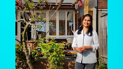 Electric blue background to make image 16x9. Vijaya Lakshmi stands in a garden setting. She wears a light grey and dark grey trousers. She smiles to camera and holds her hands together.