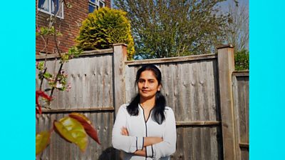 Electric blue background to make image 16x9. Vijaya Lakshmi stands in front of a fence with a little bit of a plant (about to bloom) in the foreground. She wears a light grey. She smiles to camera and has her arms folded.