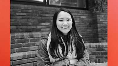 Burnt orange background to make 16 x9. Black and white photo of Vivian Young, smiling to camera. Long hair passed shoulder. Arms folded. Outside with brick wall behind her. 